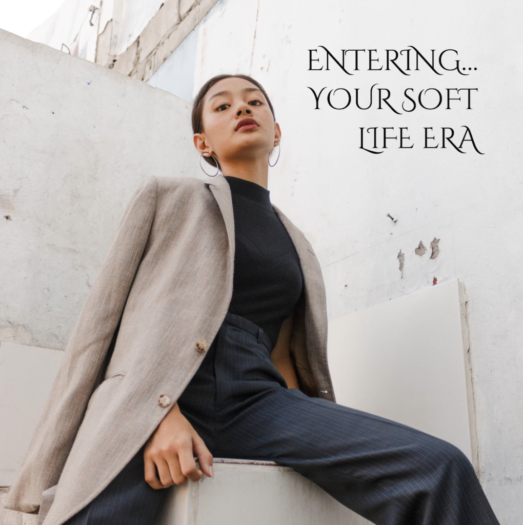 Woman wearing black pants and tan jacket sitting in front of an off-white wall